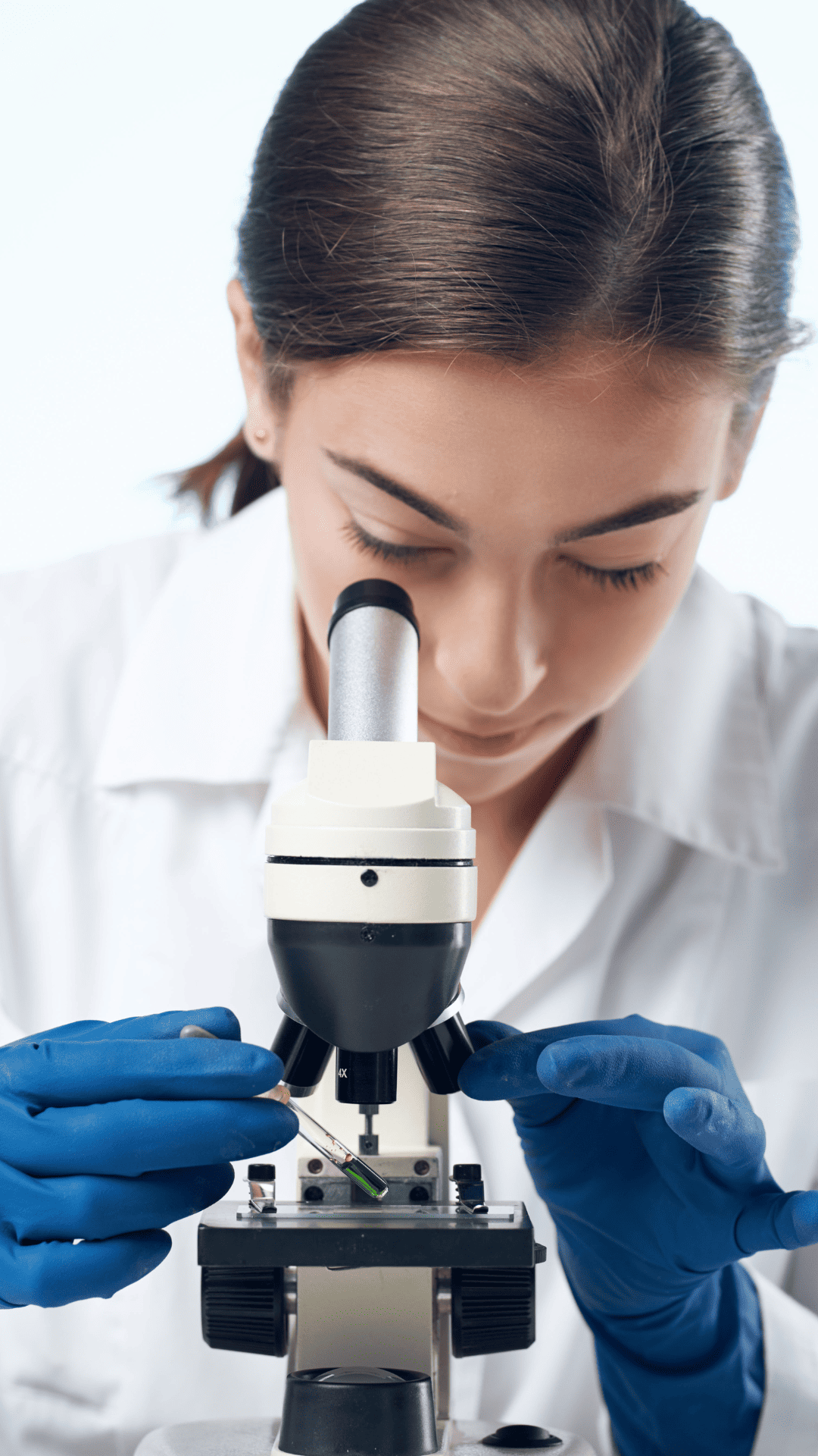 A woman in a lab coat examines a sample through a microscope in a laboratory setting.